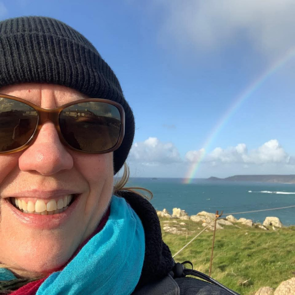 Jane Gammage wearing sunglasses and a beanie with a rainbow over the ocean in the background